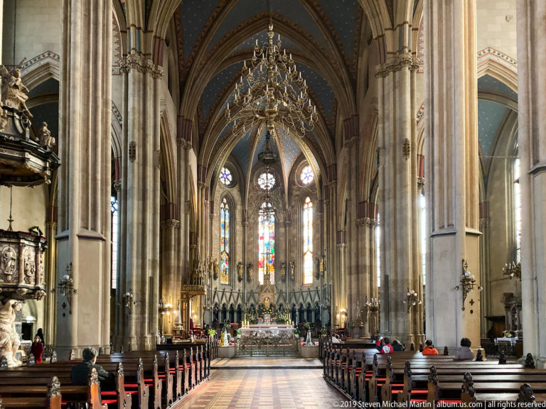 Long Gothic cathedral nave with clustered stone piers, ribbed vaults, chandelier, pews, and stained-glass windows near the sanctuary.