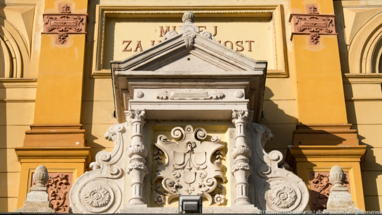 Ornate stone entrance surround with columns, scrollwork, shields, and Croatian inscription on an ochre cultural building.