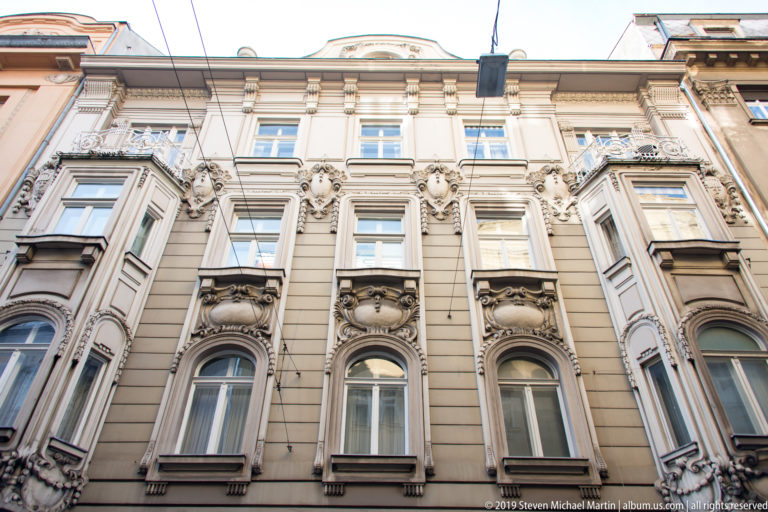 Ornate pale historic apartment façade with projecting bay windows, carved masks, floral reliefs, pilasters, and tall sash windows.