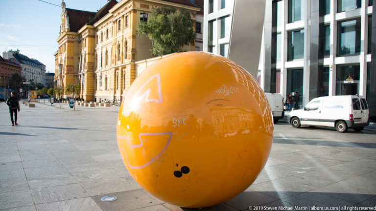 Large graffiti-marked orange sphere sculpture stands in a paved Zagreb square between historic and modern buildings.
