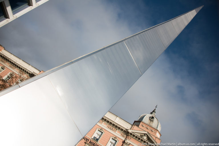 Low-angle view of a tall white tapered public sculpture rising against the sky beside an ornate historic building.