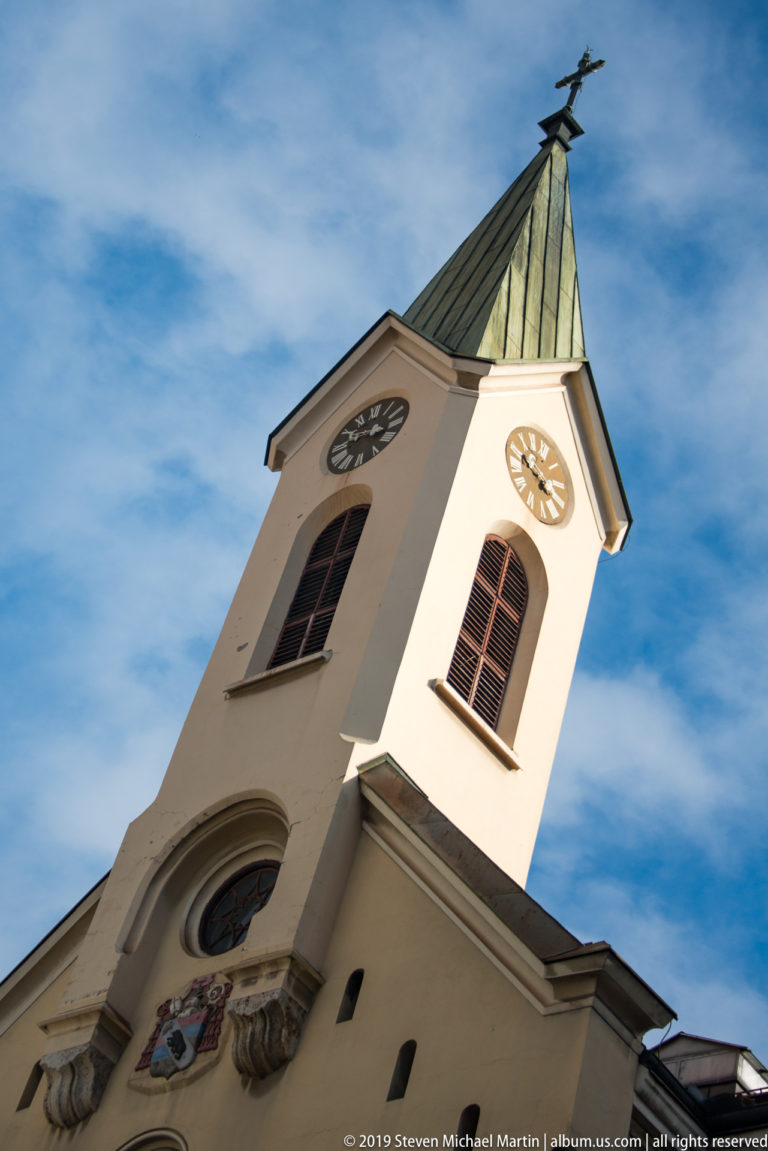 Low-angle view of a white church tower with two clocks, dark louvered openings, green metal roof, and weather vane.