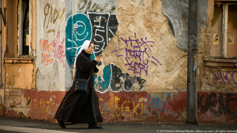 Nun in black habit walks while looking at a phone beside a cracked, graffiti-covered wall in Zagreb.