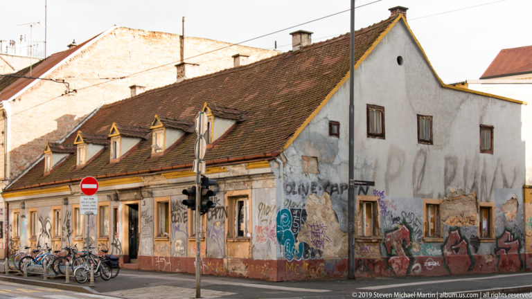 Weathered one-story Zagreb house with red-tiled roof, dormer windows, graffiti-covered walls, bicycles, and street signs.