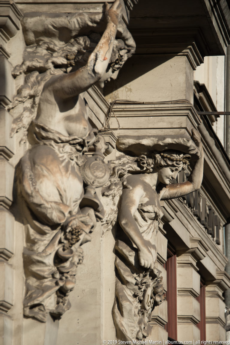 Close angled detail of sculpted female façade figures with drapery, garlands, and ornate projecting stone moldings.
