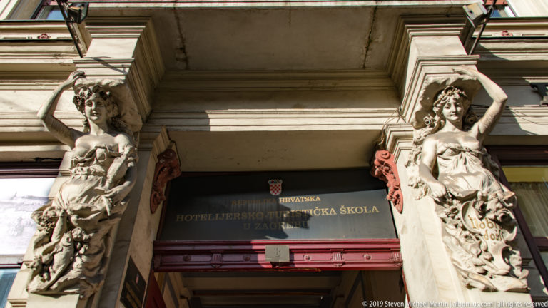 Two carved female figures flank the doorway beneath the Croatian Hospitality and Tourism School sign in Zagreb.