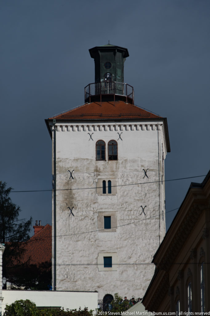 White rectangular Lotrščak Tower with small windows, red-tiled roof, iron tie plates, and enclosed lookout structure on top.
