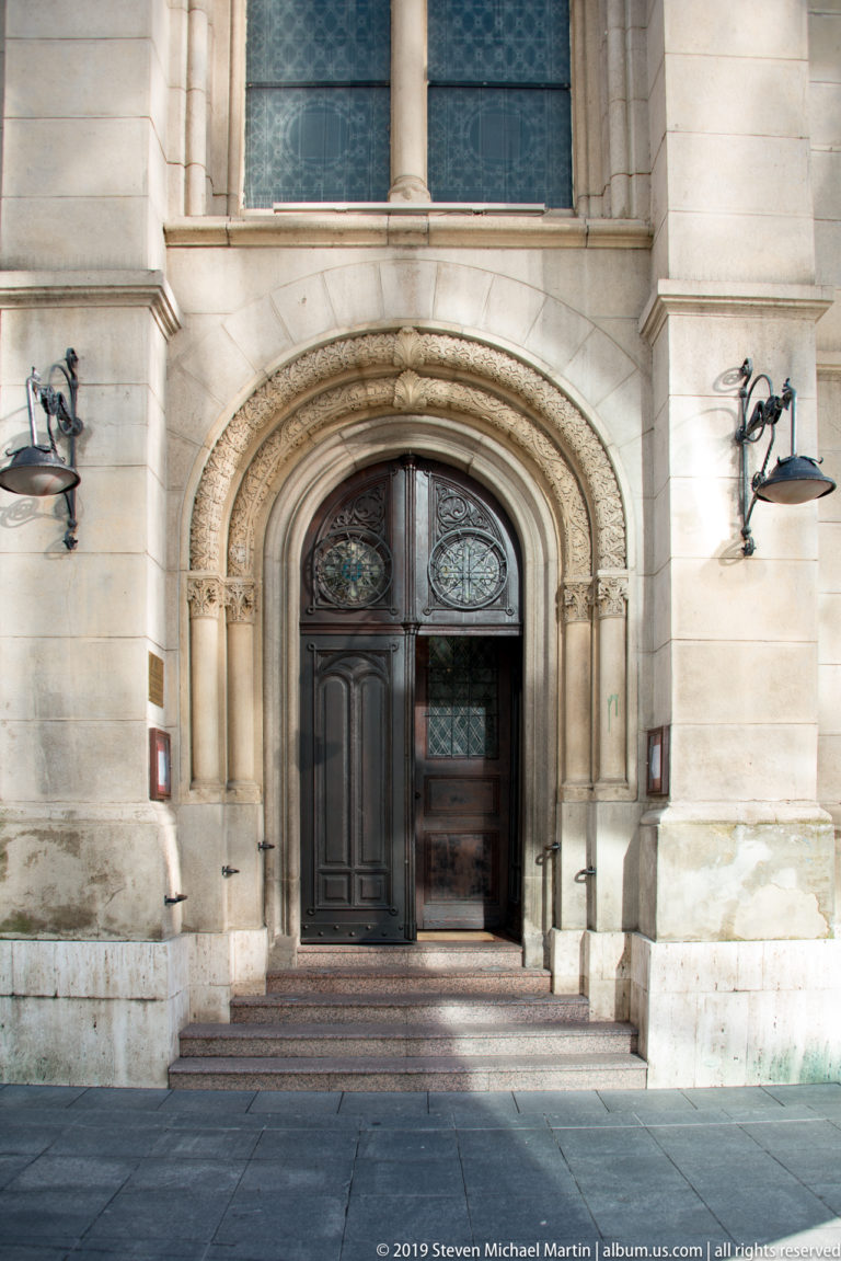 Carved round-arched stone doorway with dark wood-and-glass double doors, shallow steps, and decorative wall lamps.