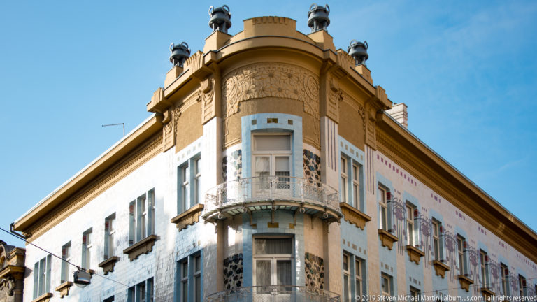 Upward view of a rounded historic corner façade with pale blue panels, floral ornament, mosaic-like decoration, and rooftop finials.