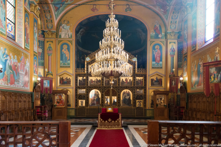 Full nave view toward a gilded iconostasis, with red carpet, wooden stalls, large chandelier, murals, and patterned floor.