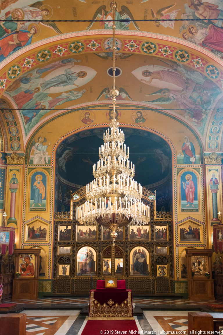 Wide symmetrical church interior with large crystal chandelier, gilded iconostasis, painted walls, and red liturgical table.