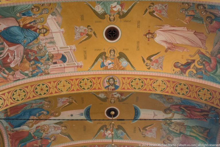 Upward view of a gold-toned Orthodox church vault painted with angels, saints, medallions, and ornamental borders.