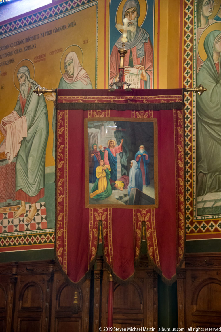 Red velvet liturgical furnishing with gold trim and small religious painting against walls covered with saint murals.