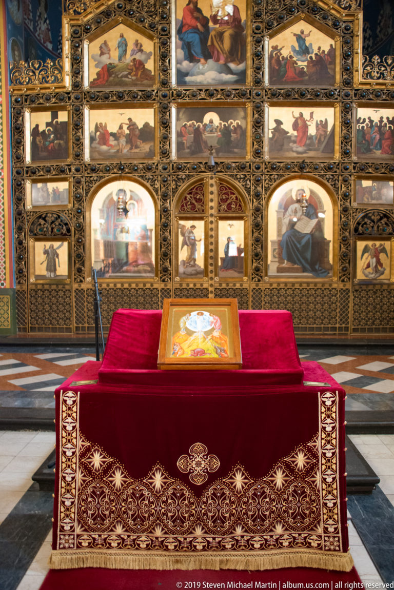 Red embroidered liturgical table with an icon stands centered before a richly gilded Orthodox iconostasis.