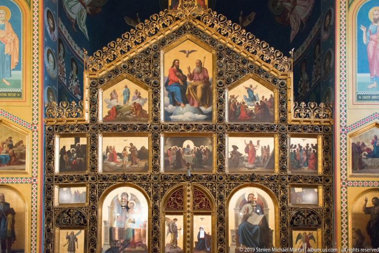 Upper portion of a gilded Orthodox iconostasis with rows of painted biblical scenes and elaborate scrolling ornament.