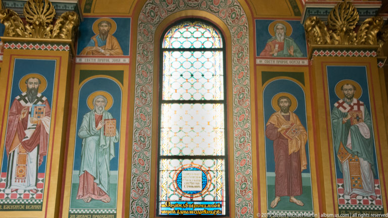 Four painted saints in tall panels flank a narrow patterned stained-glass window inside an Orthodox church.