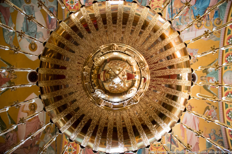 Direct upward view beneath an ornate gold and crystal chandelier, with painted church ceiling visible around its edges.
