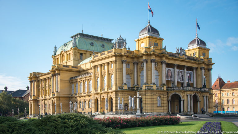Three-quarter view of Zagreb’s yellow Croatian National Theatre with columns, rooftop sculptures, corner towers, flags, and landscaped flower beds.