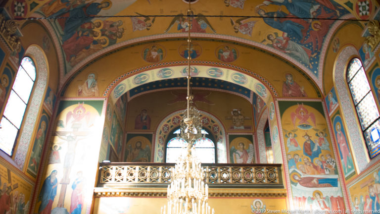 Painted Orthodox church nave with balcony, arched windows, saints, biblical scenes, and a central crystal chandelier.