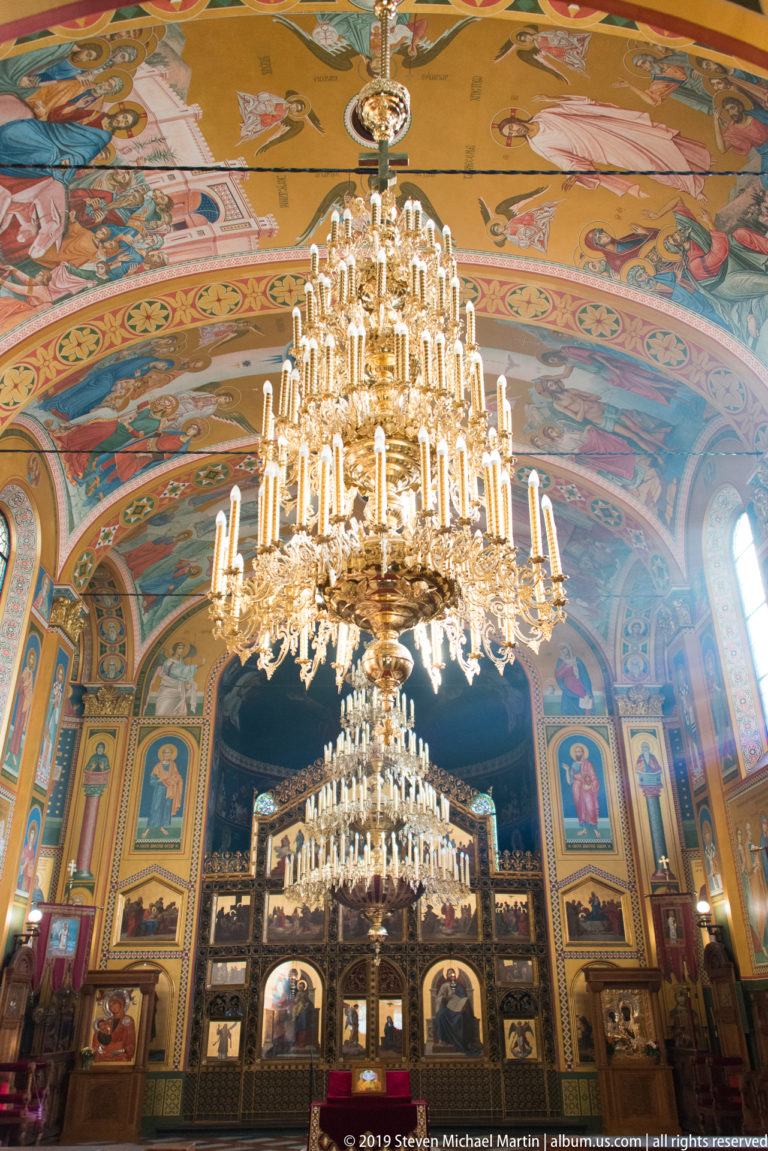 Large crystal chandelier hangs before a gilded iconostasis in a vividly painted Serbian Orthodox church interior.