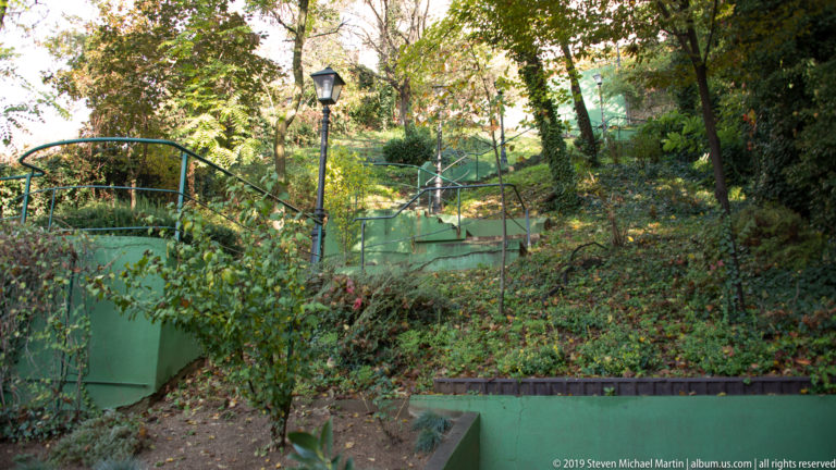 Wooded hillside path with green railings, steps, dense vegetation, and a black streetlamp in Zagreb.