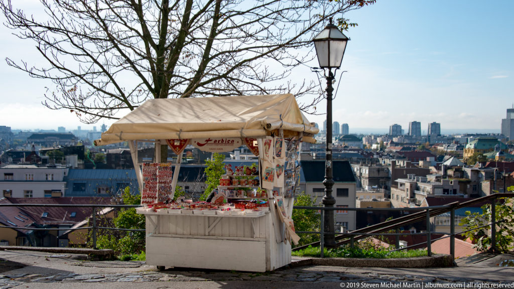 Small white souvenir kiosk with colorful merchandise beside a black streetlamp, overlooking the Zagreb cityscape.