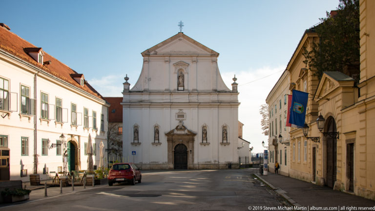 White Serbian Orthodox cathedral façade with central portal, saint niches, triangular pediment, and cross at the end of a narrow street.