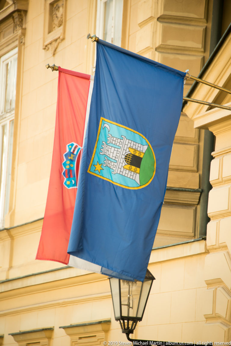 Croatian national flag and blue Zagreb city flag hang from a pale historic façade above a black wall lantern.
