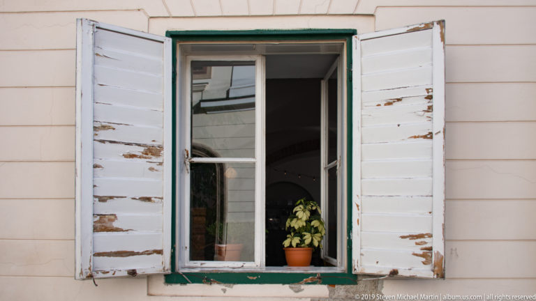Tall green-trimmed window with peeling white wooden shutters and a small potted plant on the interior sill.