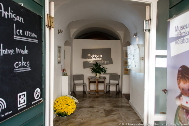 Arched museum entrance corridor with Museum of Broken Relationships sign at the far wall, open green door, café chalkboard, and yellow flowers.