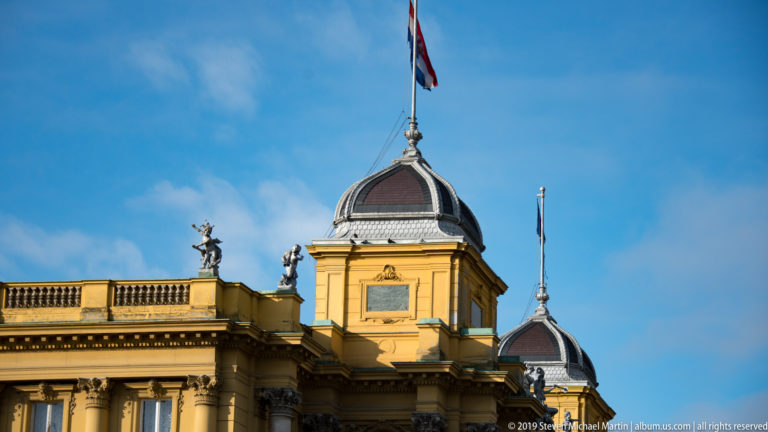 Two ribbed copper-roofed pavilions with finials and a Croatian flag above an ornate ochre historicist façade.