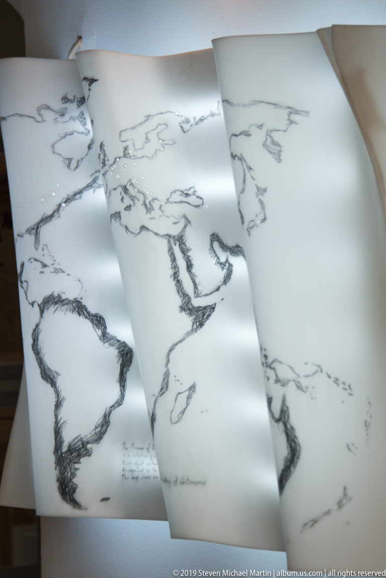 Overlapping white sheets printed or drawn with sketchy world maps, suspended or folded into a layered museum installation.