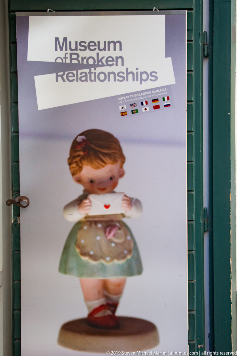 Museum of Broken Relationships poster in a green doorway with museum title, small figurine image, and a row of national flags.