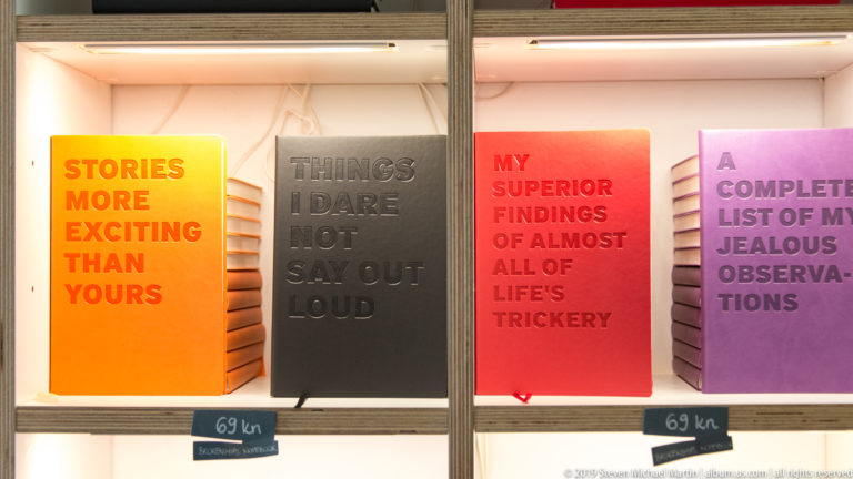 Orange, black, red, and purple notebooks on a museum shop shelf, each printed with humorous relationship-themed phrases.