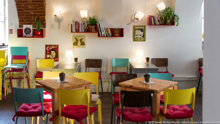 Bright café interior with mismatched colorful chairs, small wooden tables, books, plants, wall lamps, and red seat cushions.
