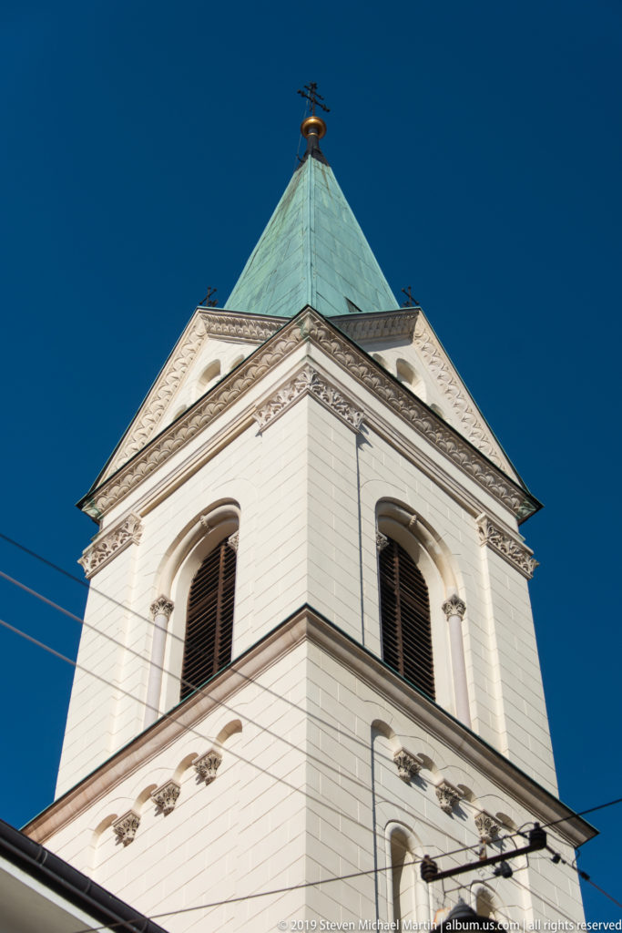 Low-angle close view of a white bell tower with arched louvered openings and a green pyramidal roof against blue sky.