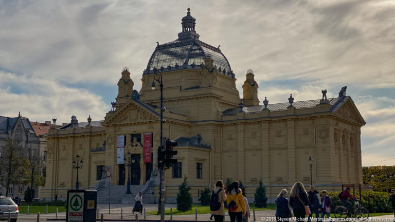 Ochre Art Pavilion in Zagreb with central glass dome, classical façade, statues, and visitors walking through the park foreground.