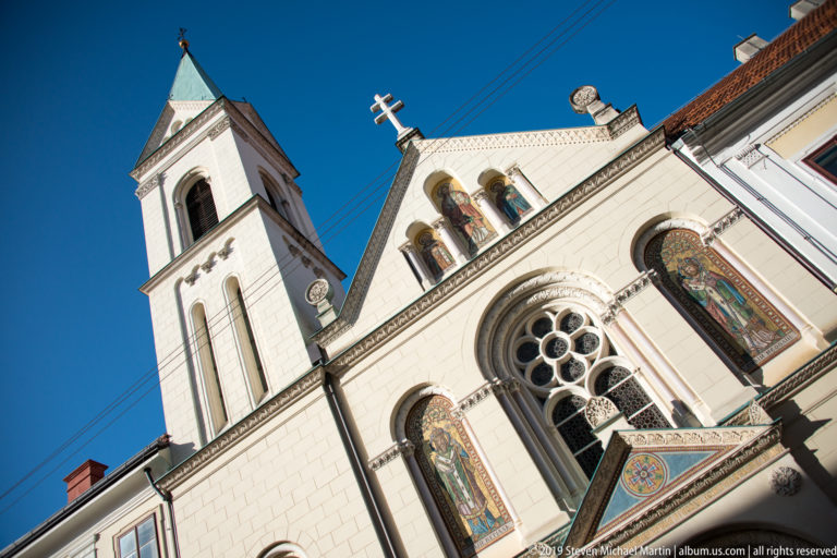 Low-angle view of pale church façade with square bell tower, round tracery window, arched openings, and colorful religious mosaics.