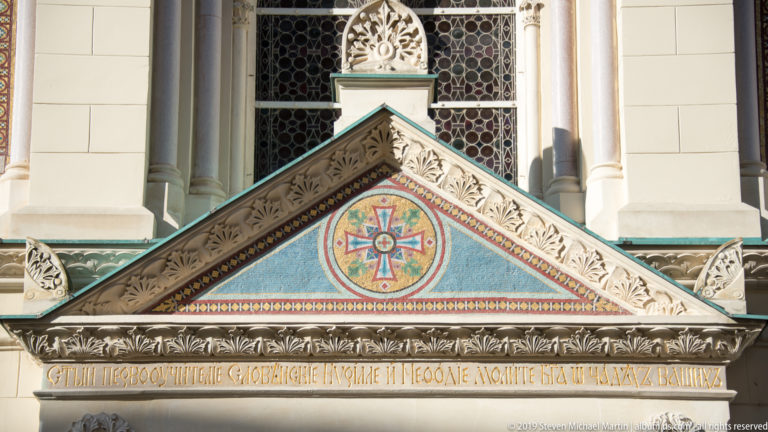 Triangular church gable with colorful cross medallion, carved stone ornament, inscription, and stained-glass window behind.