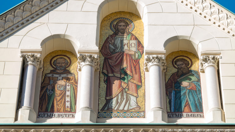 Three standing saint mosaics in arched façade niches, with gold backgrounds, halos, columns, and inscriptions below.