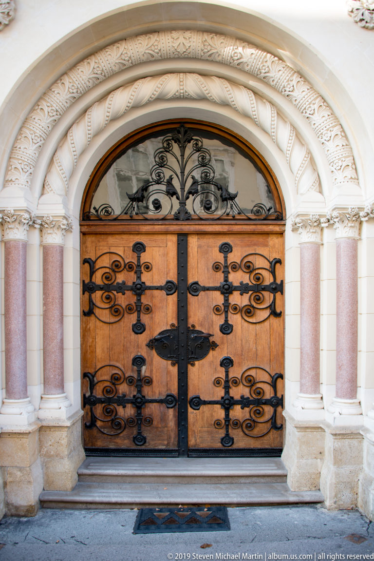 Ornate wooden double doors with black iron strap hinges set between pink columns beneath a carved round arch.