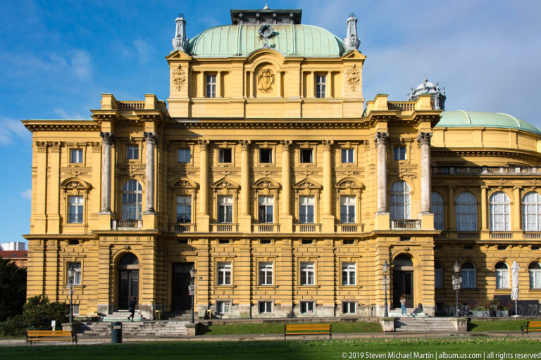 Frontal view of an ornate ochre civic building with rusticated base, arched windows, sculpted pediments, and broad green roof.
