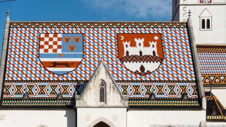 Patterned glazed-tile roof of St. Mark’s Church with two heraldic shields above pale Gothic gables.