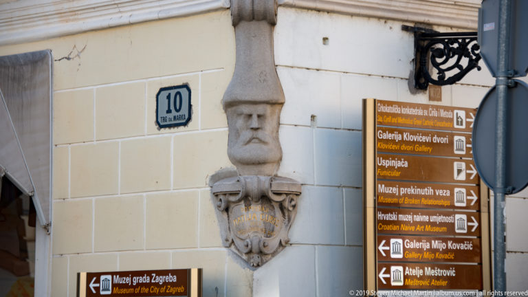 Brown directional signs for Zagreb museums and galleries mounted beside a carved stone detail on a pale Upper Town façade.