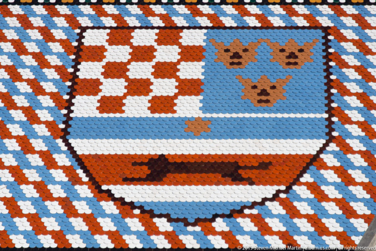 Composite heraldic shield in glazed tiles with Croatian checkerboard and blue-red fields, surrounded by patterned roof tiles.