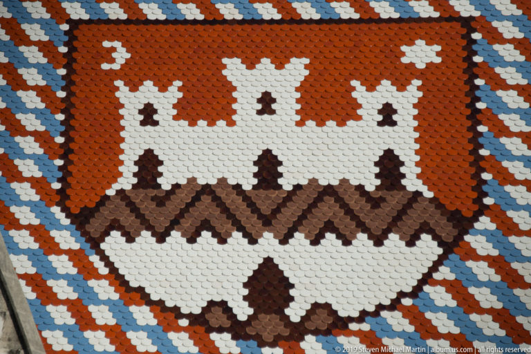 Glazed-tile Zagreb coat of arms with white three-towered city gate on red field, surrounded by multicolored roof patterns.