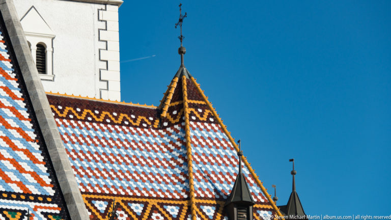 Steep gabled section of St. Mark’s Church roof with patterned glazed tiles, dark ridge caps, and a metal finial.