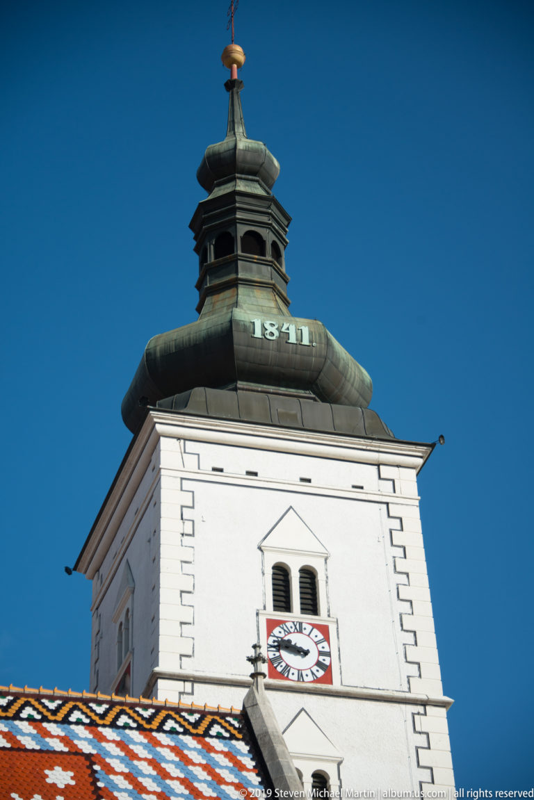 White square bell tower of St. Mark’s Church with clock face and dark metal roof above colorful glazed tiles.
