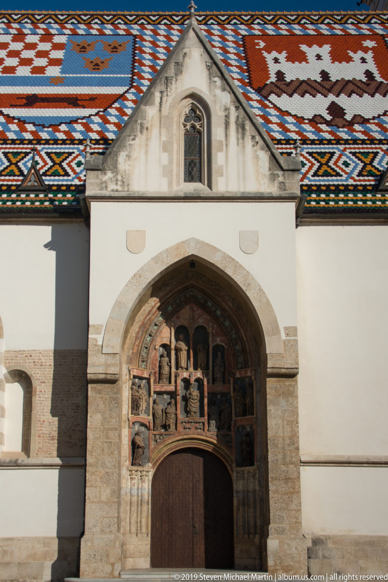 Pointed sculptured portal and wooden door of St. Mark’s Church beneath the church’s red, white, and blue tiled roof.