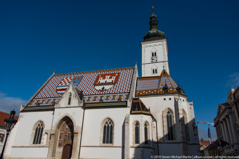 Low-angle oblique view of St. Mark’s Church showing pointed entrance, lancet windows, heraldic tiled roof, and square tower.